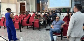 Foto de um auditório com cadeiras vermelhas organizadas em fileiras. Na frente do espaço, uma pe...