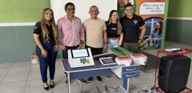 Cinco pessoas posam lado a lado em um ambiente escolar ou institucional. À frente delas há uma m...