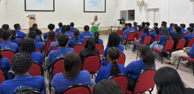 A foto mostra um auditório iluminado, com várias fileiras de cadeiras vermelhas ocupadas por ado...