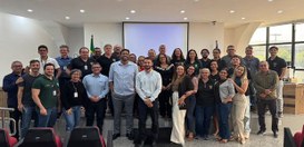 Foto de grupo em um auditório institucional. Várias pessoas estão em pé, organizadas em filas, p...