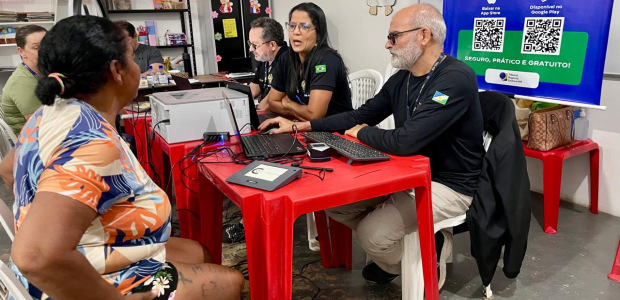 TRE-RR- Participa da segunda edição do Mutirão PopRuaJud Uma mulher sentada de frente conversa com três pessoas que trabalham em uma mesa vermelha com la...