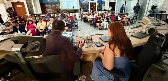 Fotografia de uma reunião em auditório. Em primeiro plano, duas pessoas estão sentadas à mesa pr...