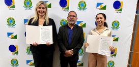 Três pessoas estão em pé diante de um painel institucional com logotipos do Tribunal Regional El...