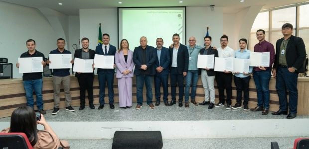 TRE-RR - Posse novos servidores concurso unificado A imagem mostra um grupo de 14 pessoas em pé, lado a lado, em um auditório. No fundo, há uma tel...