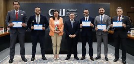 A imagem mostra um grupo de sete pessoas em pé, lado a lado. Elas estão posicionadas no centro d...