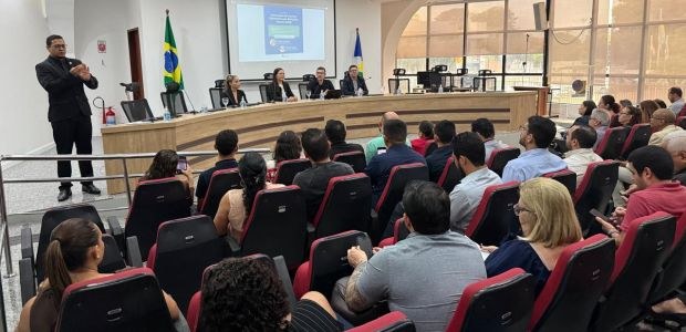 Fotografia de uma palestra realizada em um auditório. Dezenas de pessoas estão sentadas em poltr...