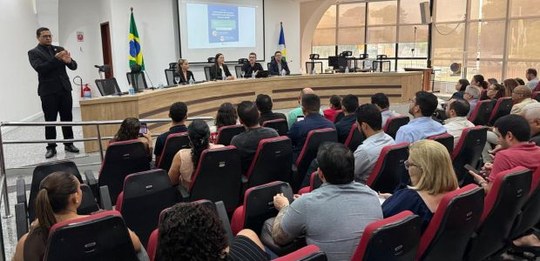 Fotografia de uma palestra realizada em um auditório. Dezenas de pessoas estão sentadas em poltr...