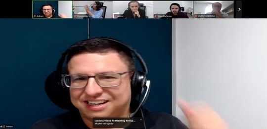 Tela de videoconferência com várias janelas. Na parte central, uma pessoa usa fones de ouvido co...