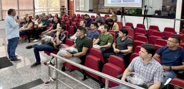 A imagem mostra um auditório com cadeiras vermelhas ocupadas por servidores e servidoras atentos...