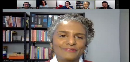 Imagem de uma videochamada. Em destaque, uma pessoa fala diante da câmera, com fundo de uma esta...