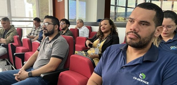 Foto de uma sala de auditório com cadeiras vermelhas em fileiras. Várias pessoas adultas estão s...