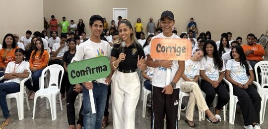 Imagem em ambiente fechado com várias pessoas sentadas em cadeiras, organizadas como plateia. À ...