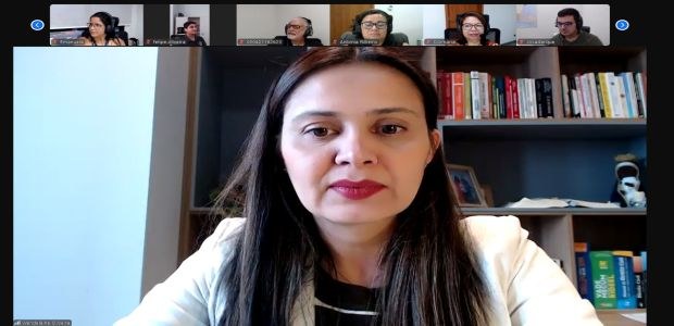A imagem mostra uma reunião virtual. No centro, em destaque, está uma mulher de pele clara e cab...