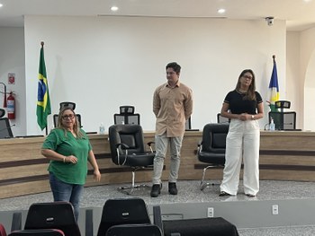 Foto de um auditório com um palco à frente. Três pessoas adultas estão em pé no palco, diante de...