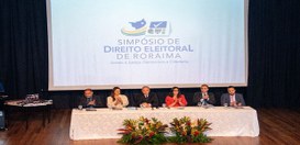 Ambiente de auditório com mesa longa coberta por toalha branca no palco, onde várias pessoas est...