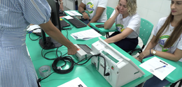 Em uma sala com mesas verdes, várias pessoas sentadas participam de uma atividade com equipament...