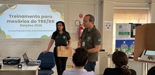 Foto de um ambiente interno de treinamento com participantes sentados e duas pessoas em pé à fre...