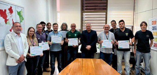 Fotografia de um grupo de cerca de doze pessoas, em pé, lado a lado, dentro de uma sala de reuni...
