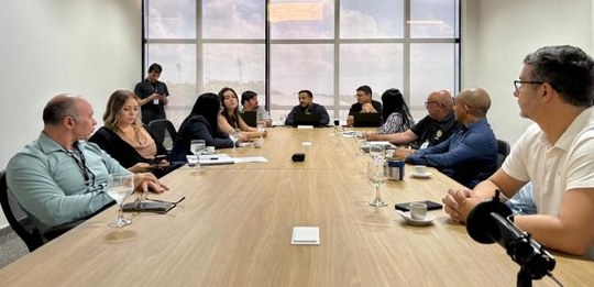 A foto mostra uma reunião em uma sala de conferências. Várias pessoas estão sentadas ao redor de...