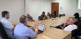 Sala de reuniões com mesa ampla de madeira em formato retangular. Oito pessoas estão sentadas ao...