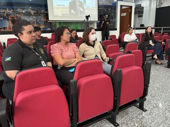 Um grupo de pessoas está sentado em poltronas vermelhas de um auditório, assistindo a uma aprese...