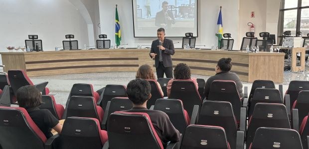 Um homem em pé, de terno escuro, fala ao microfone em um auditório com cadeiras vermelhas e pret...