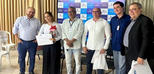 Seis pessoas estão em pé, lado a lado, posando para uma foto em um ambiente interno. Atrás delas...