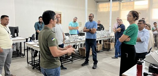 Foto em ambiente de treinamento técnico. Um grupo de pessoas está reunido em uma sala com mesas,...