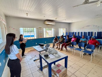 Em uma sala de aula, um grupo de pessoas está sentado em cadeiras azuis, acompanhando uma aprese...