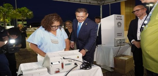 Em um ambiente externo à noite, uma urna eletrônica está sobre uma mesa branca. Ao redor, várias...