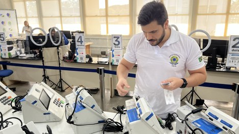 Pessoa com camisa da Justiça Eleitoral manuseia materiais ao lado de várias urnas eletrônicas, e...