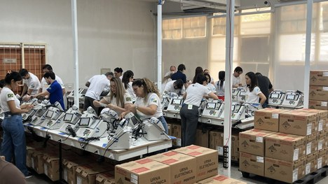Pessoa com camisa da Justiça Eleitoral manuseia materiais ao lado de várias urnas eletrônicas, e...