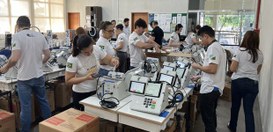 Pessoas em uma sala organizam e preparam diversas urnas eletrônicas dispostas sobre mesas, com c...