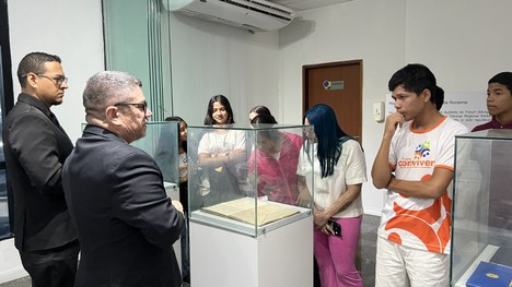 Grupo de pessoas em um ambiente interno observa um documento exposto dentro de uma vitrine de vi...