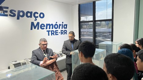Grupo de pessoas em um ambiente interno observa um documento exposto dentro de uma vitrine de vi...