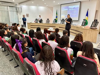 Em um auditório, uma servidora do TRE-RR fala ao microfone para um grupo de estudantes sentados....