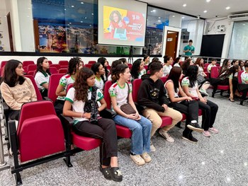 Em um auditório, uma servidora do TRE-RR fala ao microfone para um grupo de estudantes sentados....
