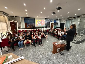 Em um auditório, uma servidora do TRE-RR fala ao microfone para um grupo de estudantes sentados....