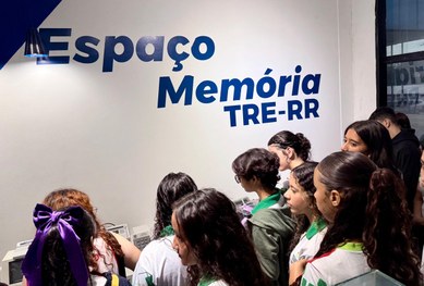 Em um auditório, uma servidora do TRE-RR fala ao microfone para um grupo de estudantes sentados....