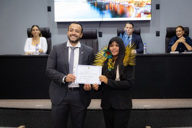 A foto mostra uma jovem indígena, vestindo traje social preto e um cocar de penas coloridas nos ...