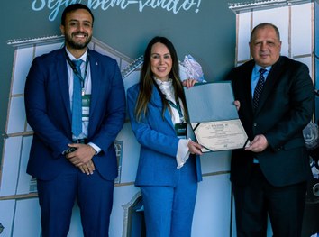 Na imagem, uma mulher de cabelos longos e lisos, vestindo blazer azul e blusa branca, segura um ...