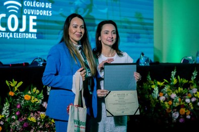Na imagem, uma mulher de cabelos longos e lisos, vestindo blazer azul e blusa branca, segura um ...