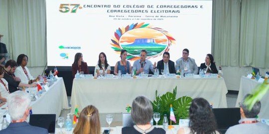 A foto mostra uma reunião formal com várias pessoas sentadas ao redor de mesas em formato de “U”...
