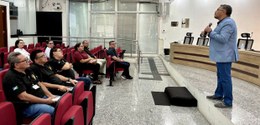 Homem em pé fala ao microfone em um auditório para um pequeno grupo de pessoas sentadas em cadei...