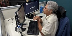 Uma pessoa de cabelos grisalhos, sentada em frente a dois monitores de computador em um escritór...