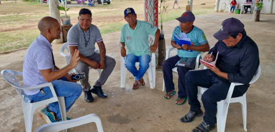 Cinco homens estão sentados em cadeiras plásticas brancas, em círculo, conversando ao ar livre. ...