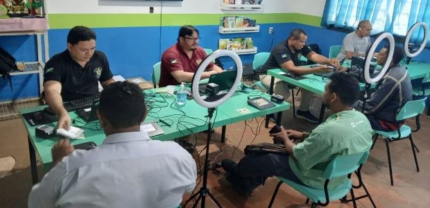A foto mostra uma sala com várias mesas verdes onde homens estão sentados, realizando atendiment...