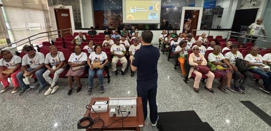A foto mostra um grupo de pessoas idosas sentadas em um auditório, assistindo a uma apresentação...