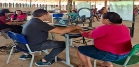 A foto mostra uma mulher sendo atendida por um servidor em uma mesa com notebook e equipamentos ...