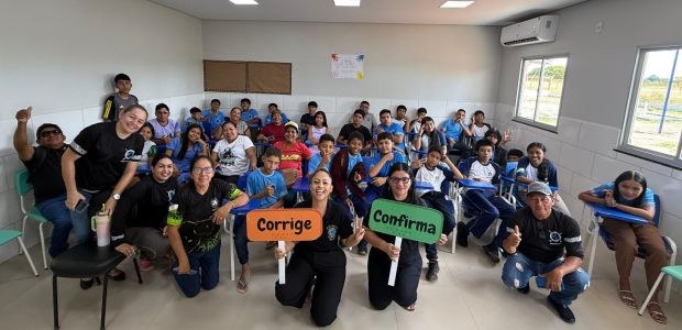 TRE-RR - Projeto “Meu Primeiro Voto” Grupo de estudantes e adultos posa sorridente dentro de uma sala de aula iluminada. Na frente, d...